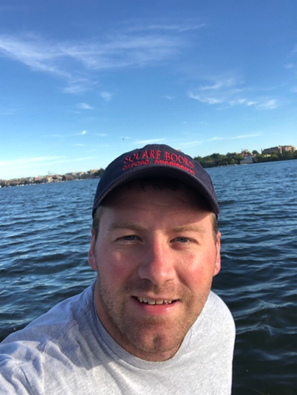 John Strauser is wearing a baseball cap and smiling at the camera. He is standing in front of a lake.